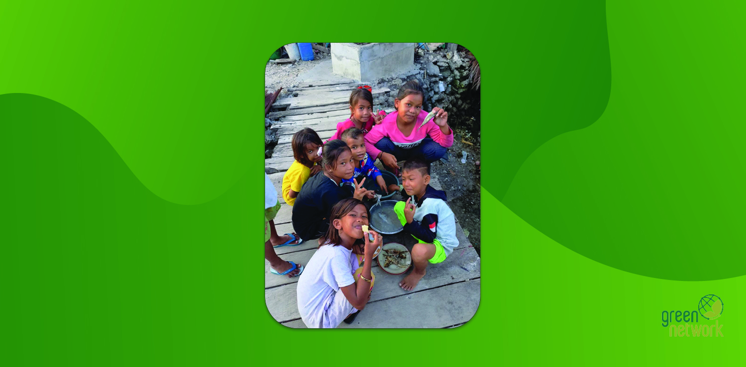 a group of children sitting around two buckets of fresh fish and a plate of fried fish, looking at the camera