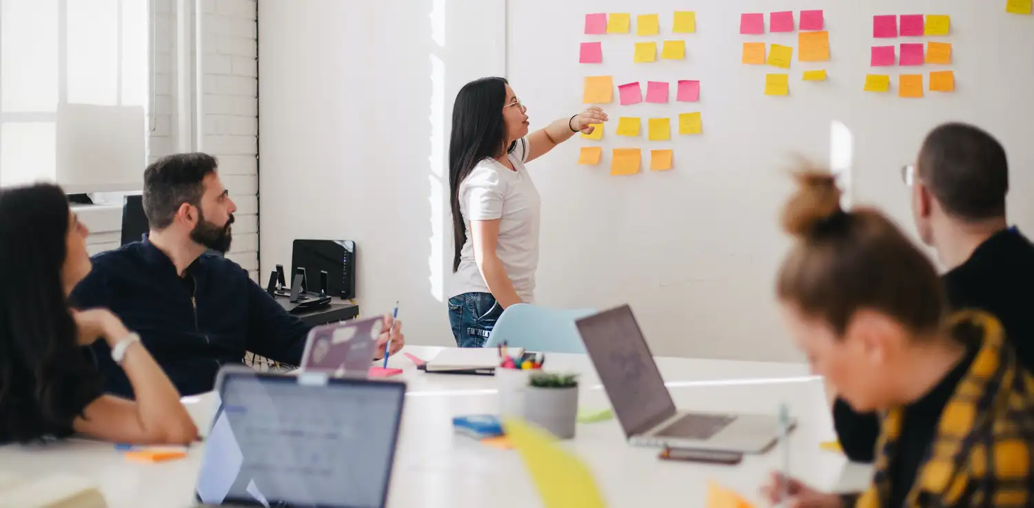 five people in a meeting, four are seated and one is explaining based on the sticky notes on the wall