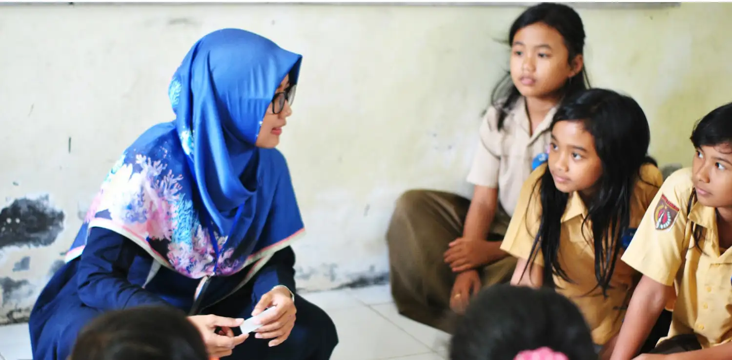 female teacher in hijab sitting cross-legged on the floor with a group of elementary school students