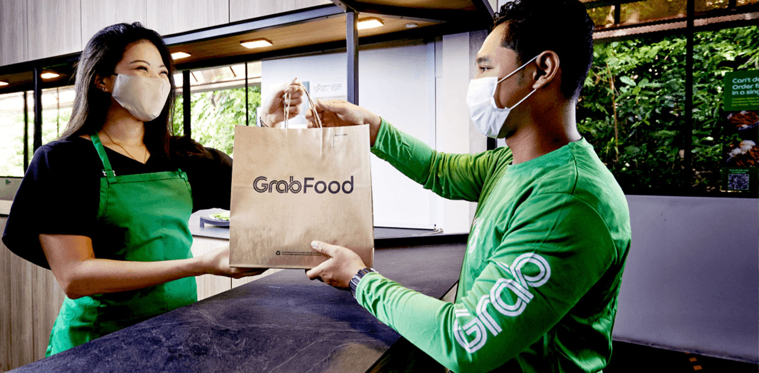 A young woman (food merchant) wearing blackshirt and green apron smiles while handing out a craft-paper bag with "GrabFood" written to a man wearing greenshirt (Grab's driver)