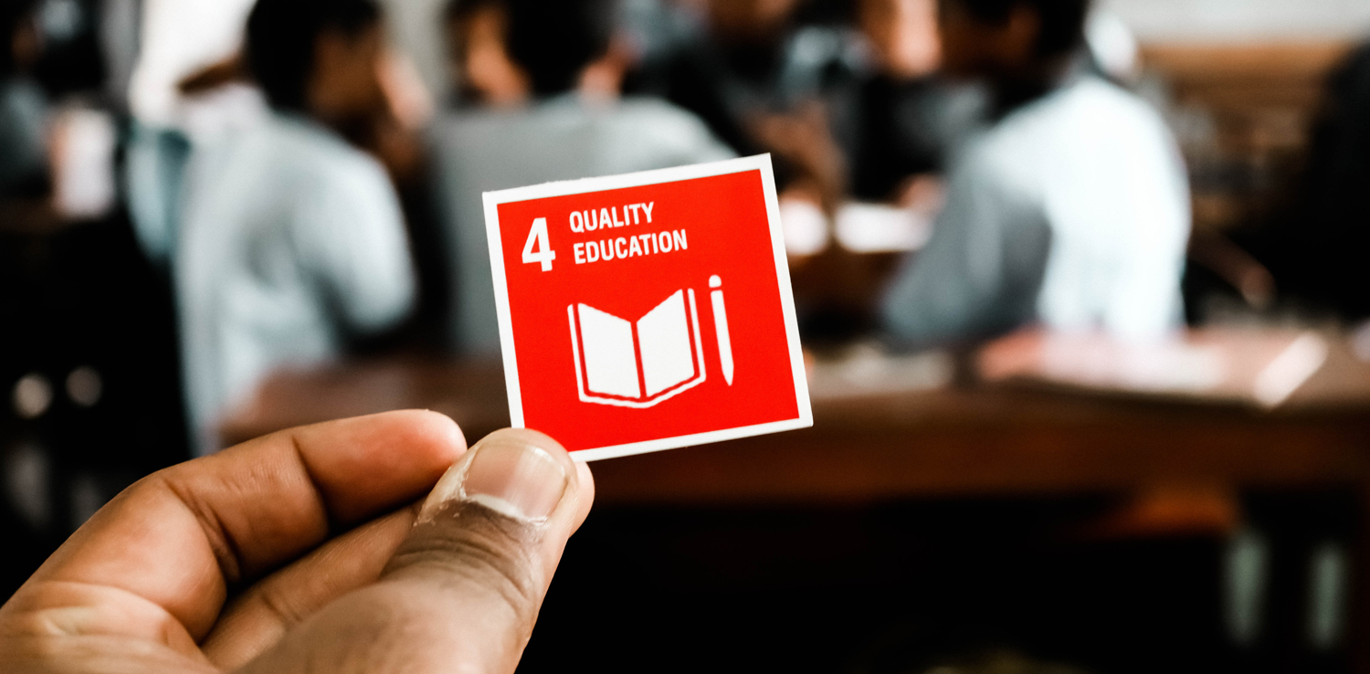 a hand holding a small red sticker printed with '4 quality education' in a classroom