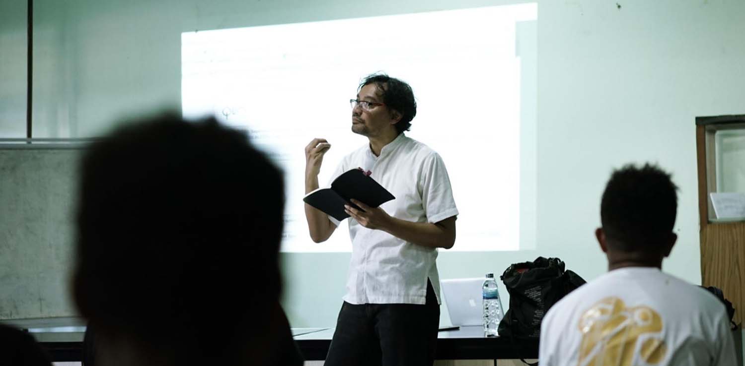 a man in white shirt holding a book speaking in front of a lecture room