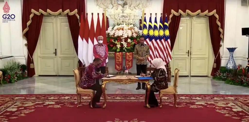 Ida Fauziyah and Dato’ Sri M. Saravanan sitting down and signing a Memorandum of Understanding on the Placement and Protection of Indonesian Migrant Workers in the Domestic Sector in Malaysia with President Joko Widodo and Prime Minister Dato’ Sri Ismail Sabri Yaakob standing behind them in front of Indonesian and Malaysian flags