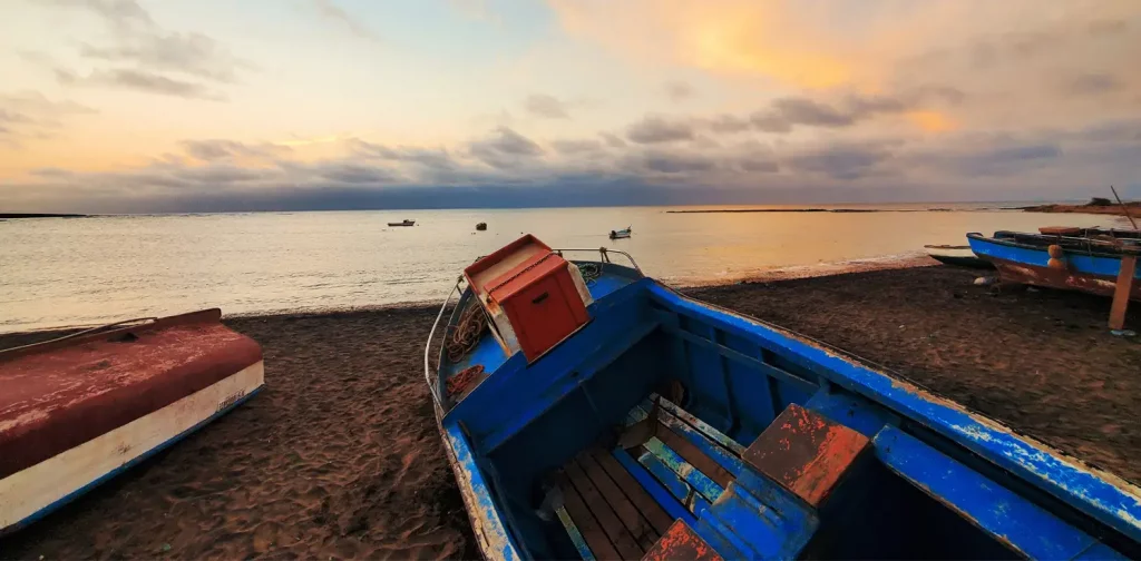 fisher boats stationed on the shore, facing the yellow-hued ocean, clouds, and blue sky