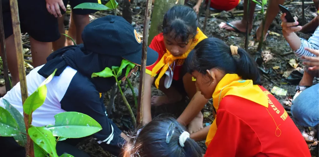 A young adult from we day ugm and three elementary schoolers planting mangrove seed. Photo is taken from above and shows only their heads and backs.
