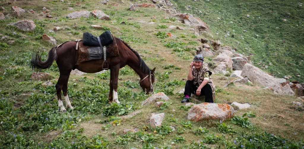 an adult woman is sitting beside a horse that is eating at a mountain’s slope