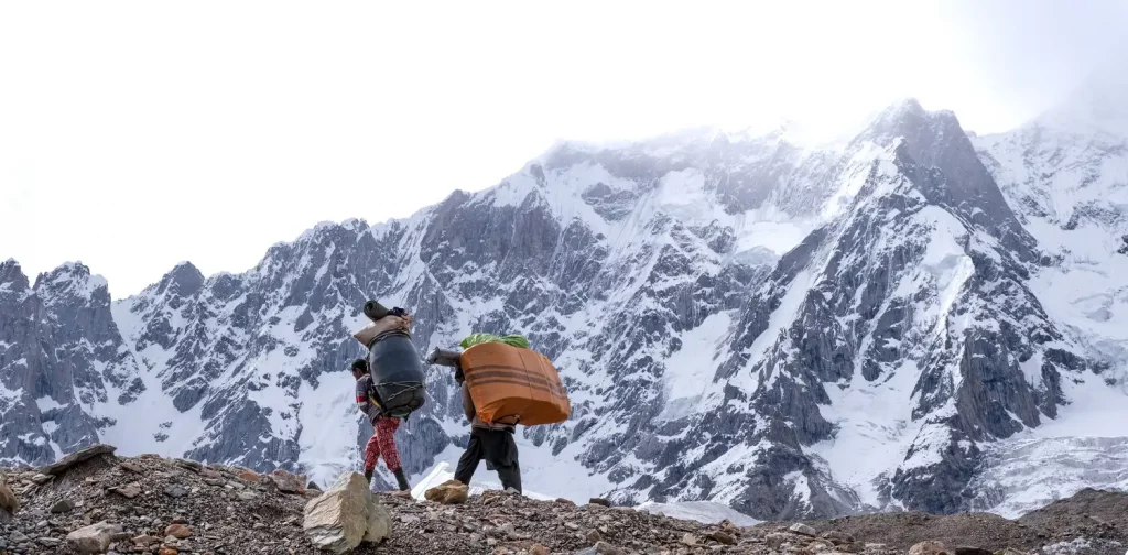two people carrying load on their backs at the baltoro glacier with icy mountain in the background