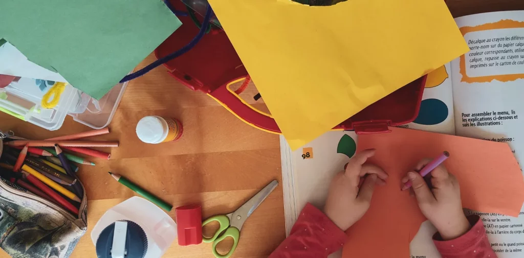 a child is in the middle of writing something on red paper, on a desk that is full of colorful papers and stationeries.