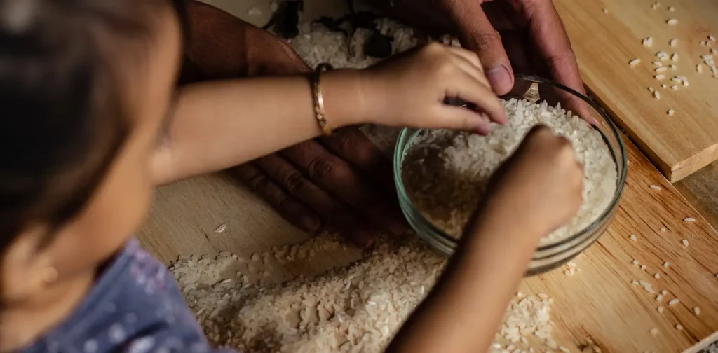 an over-the-shoulder shot of a kid curiously touching a bowl of rice grains, with a pair of adult hands holding the bowl