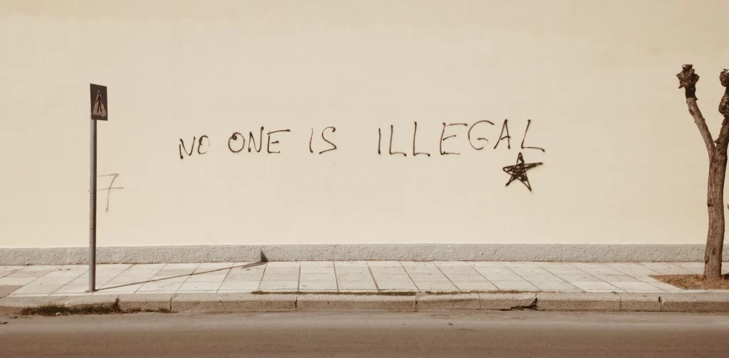 a writing that says “No one is illegal” written across a white wall against a sidewalk
