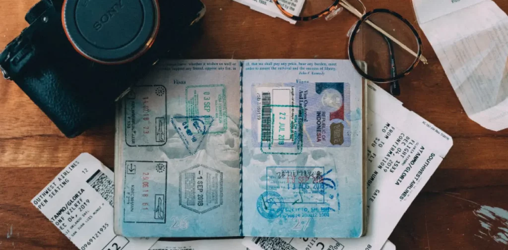 an open passport, a camera, a glasses, and few receipts are scattered on a wooden table