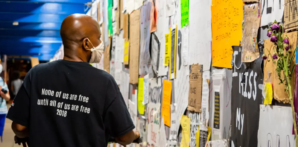 the back of a black man in black crew neck t-shirt standing beside yellow and white wall full of slogans