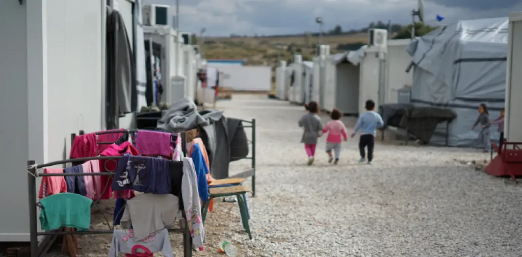 a refugee camp with three children, clothes hanging, and tents.