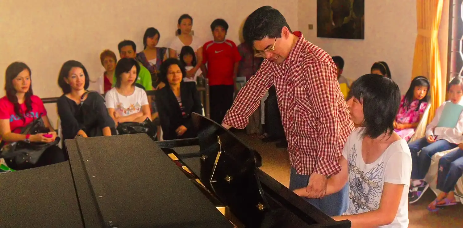 a man, Ananda Sukarlan, is teaching piano to a kid with children and parents watching in the background