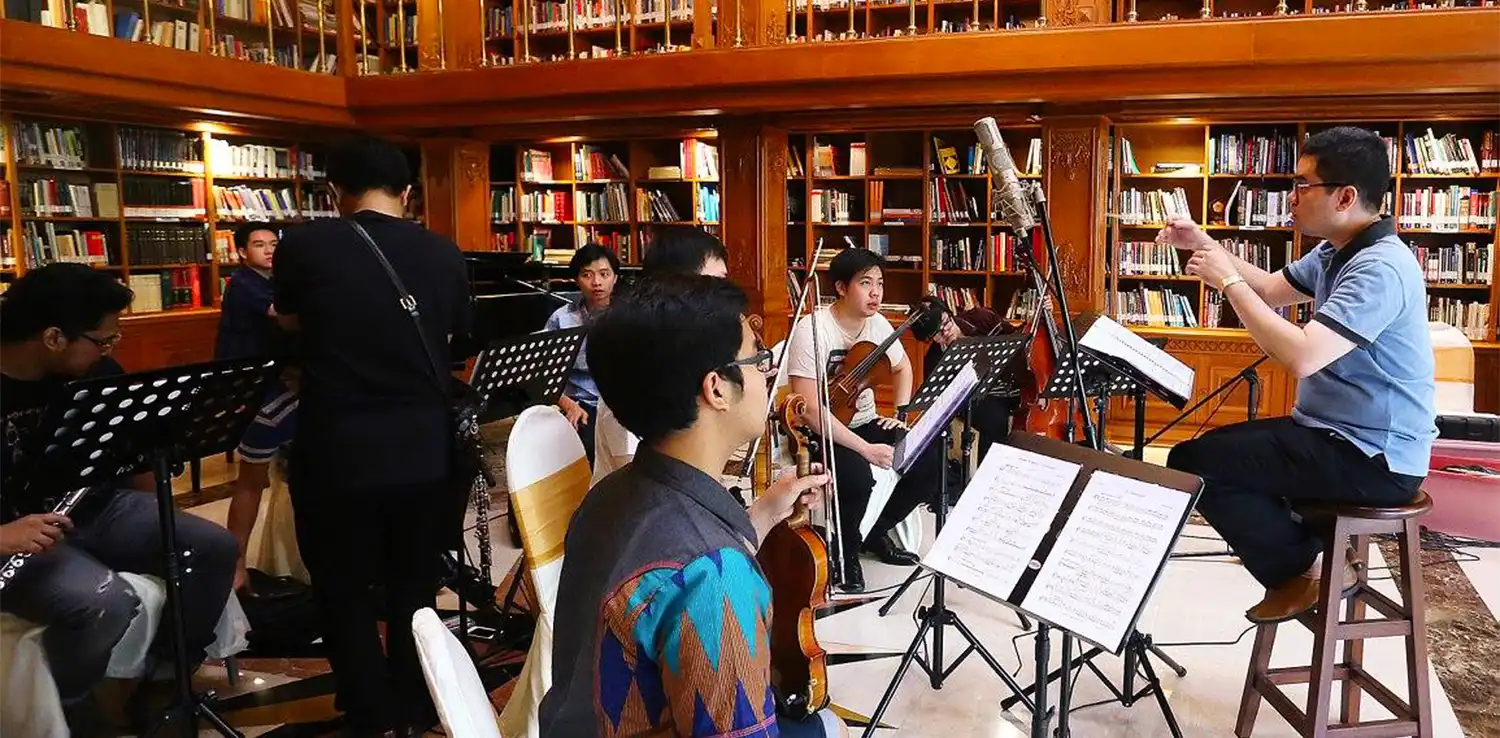 a man, Ananda Sukarlan, sitting in front of an orchestra of youth with different musical instruments 