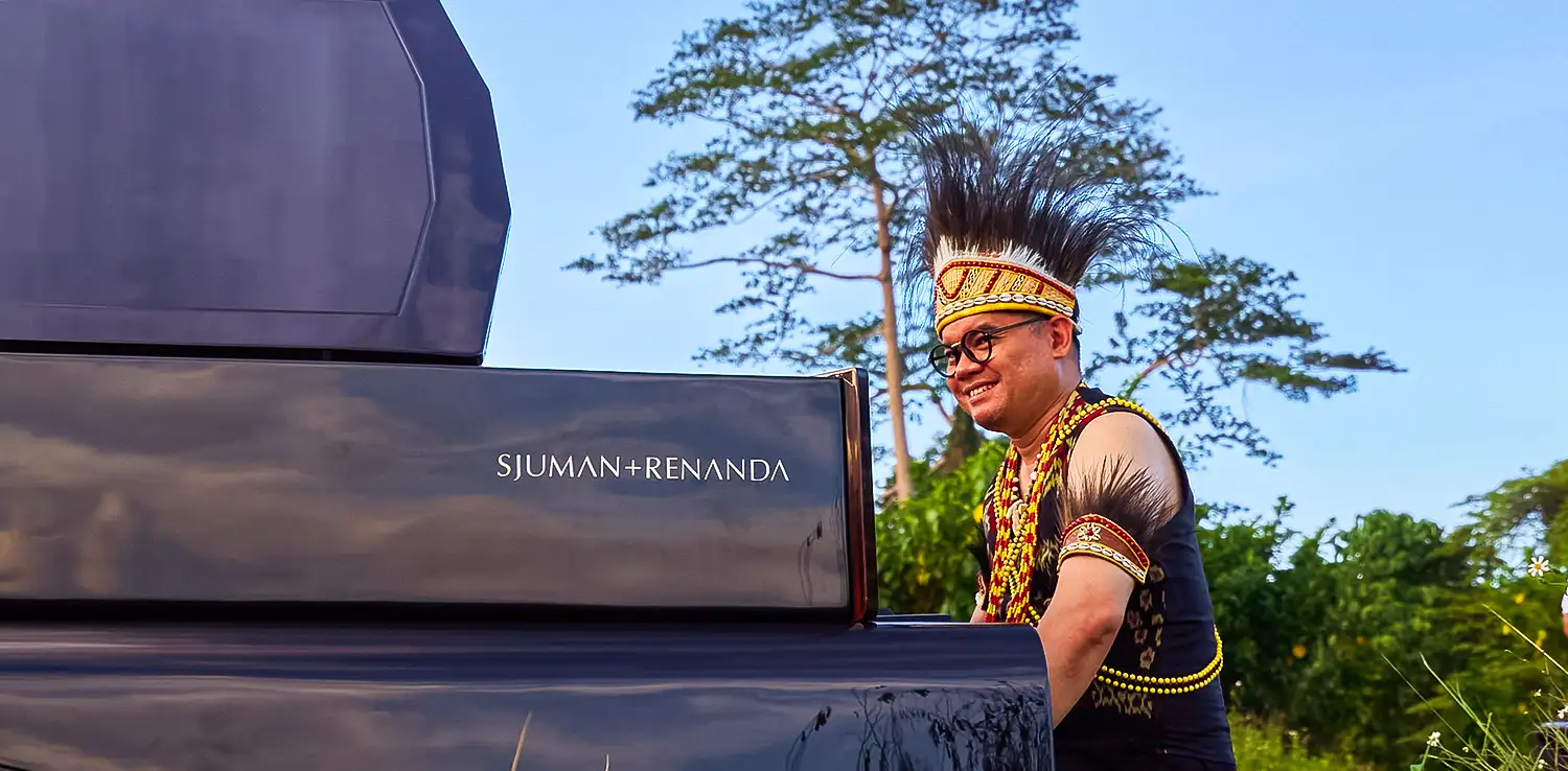 a man, Ananda Sukarlan, sitting at a black piano wearing the traditional clothes from Kalimantan