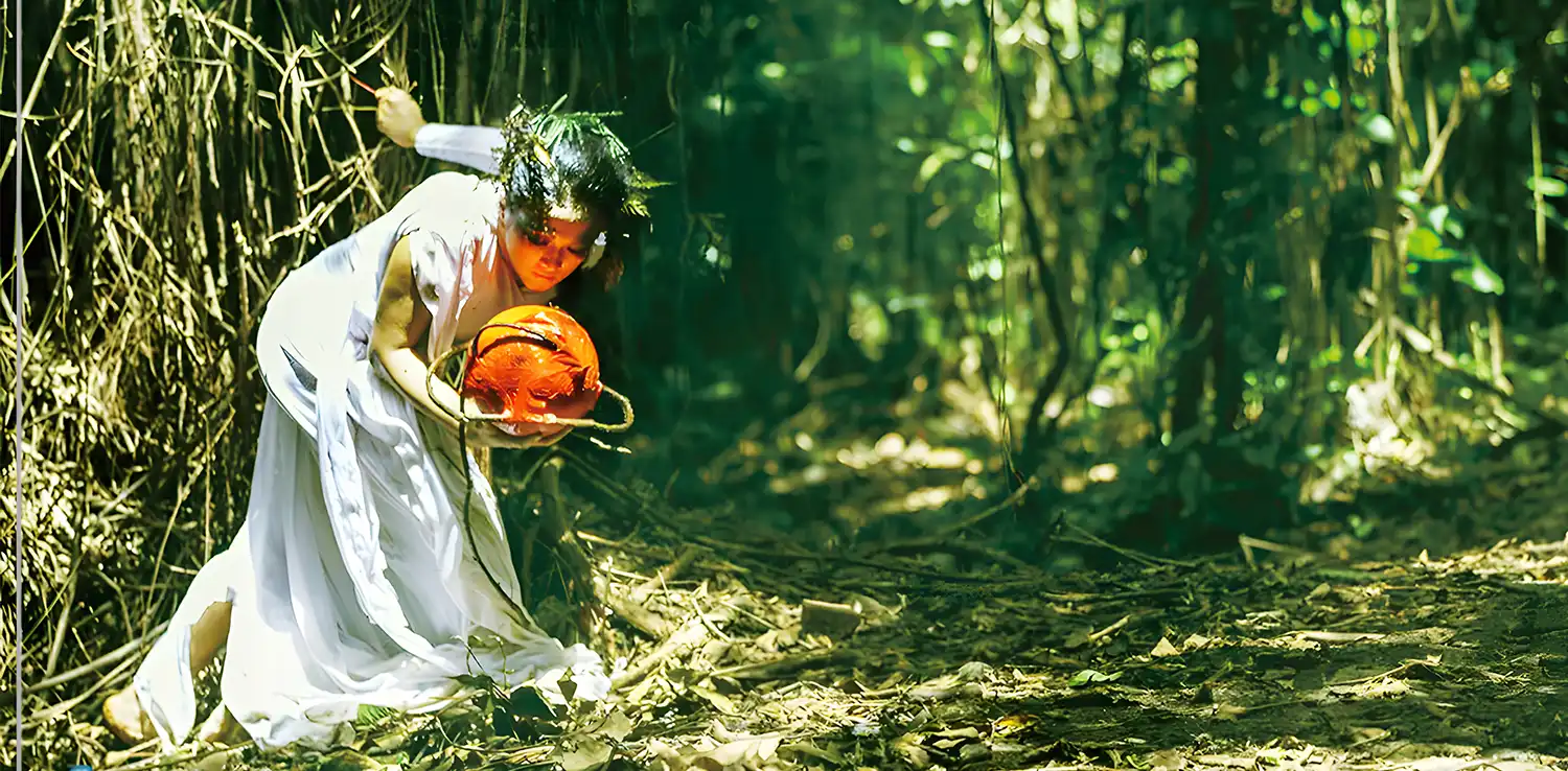 a woman, Lena Guslina, dancing in the middle of a forest wearing a white gown and holding an orange ball as a property.