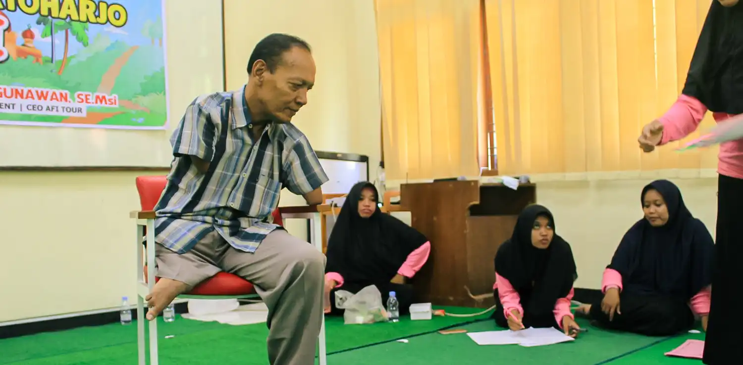 a man with phocomelia sitting on a chair on green-carpeted floor with three girls in hijab on the floor nearby