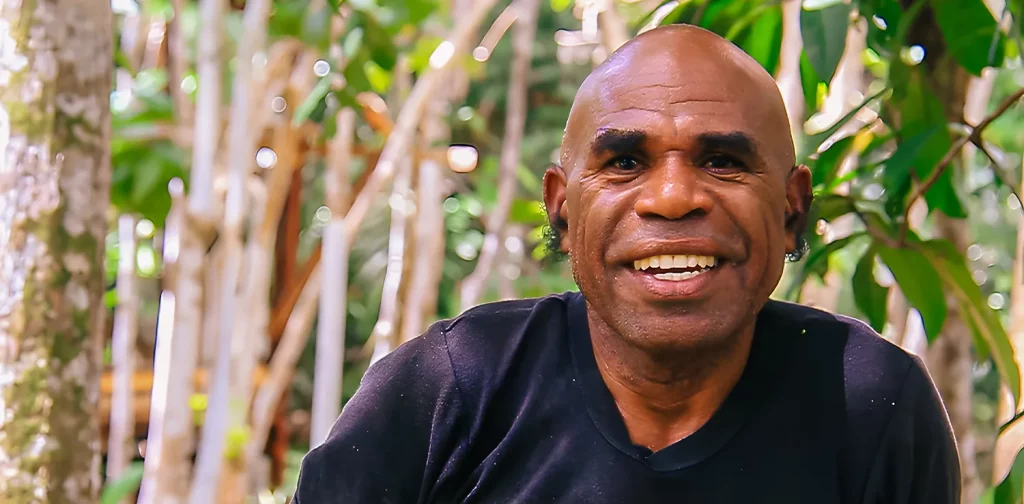 a bald man, alex waisimon, smiling at the camera with a forest in the background