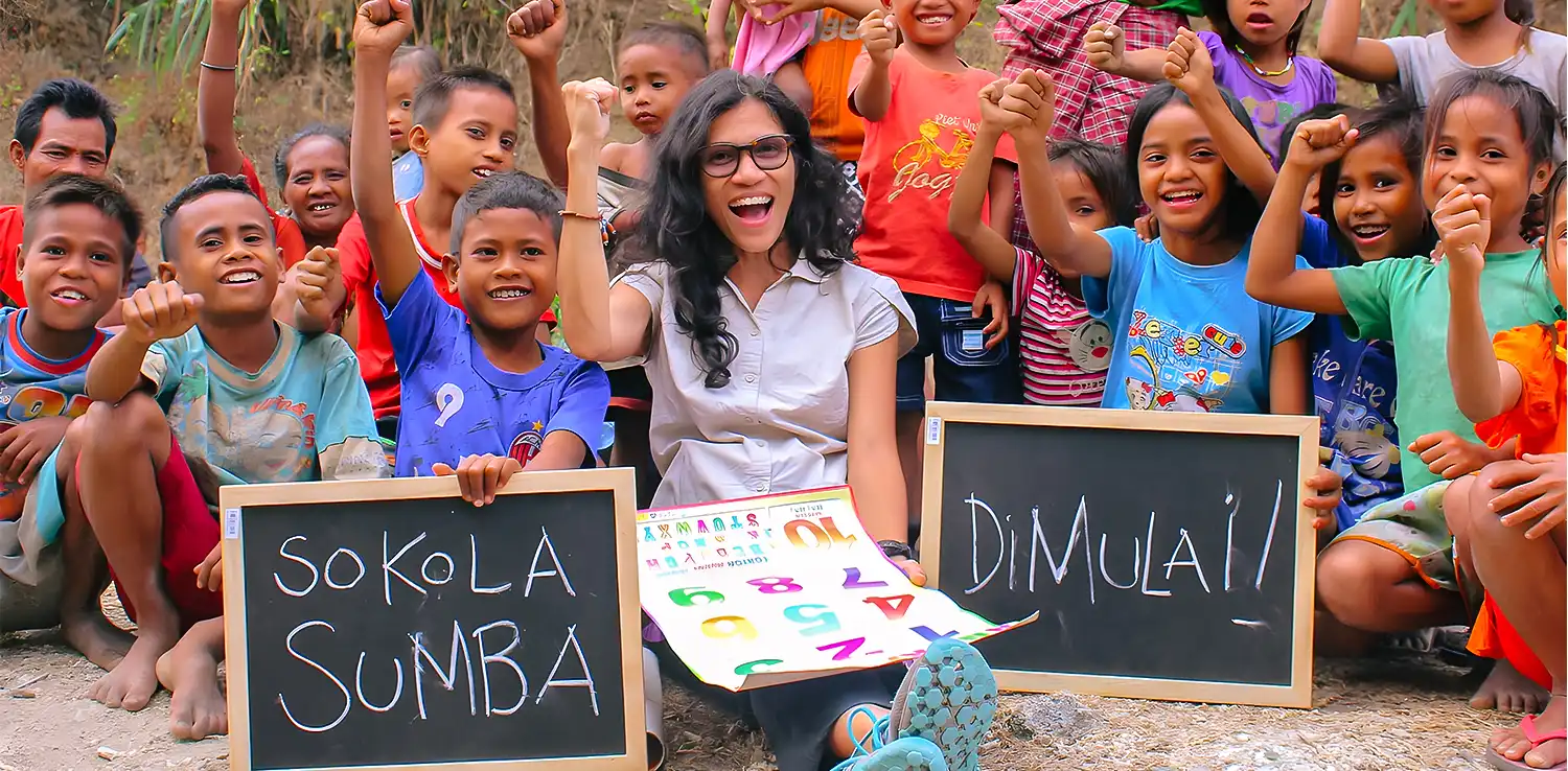 a woman, butet manurung, and children looking to the camera with boards saying ‘sokola sumba’ and ‘begins’