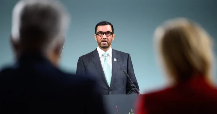 a man in suit and glasses speaking at the podium with blurry audience in the foreground