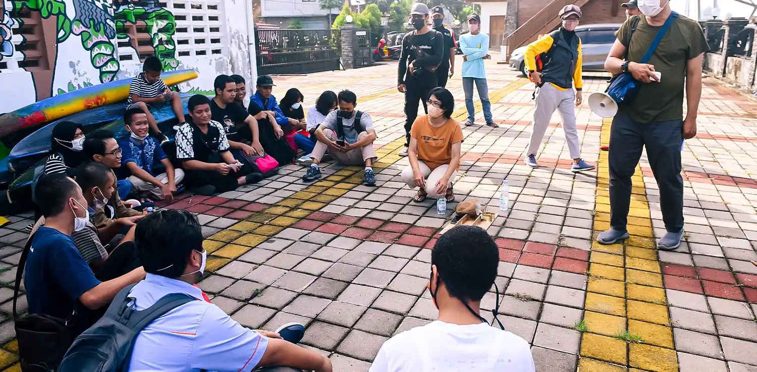 a group of people are sitting on colorful concrete blocks while paying attention to the woman, Indah Darmastuti, in the middle. A few other people are standing scattered around the group.