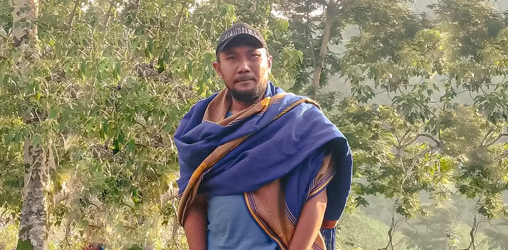 a man, Kristopel Bili, wearing a cap, t-shirt, and a blue scarf