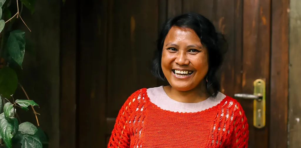 a woman, Lian Gogali, is wearing a red knitted sweater on top of white outfit while standing in front of a door.