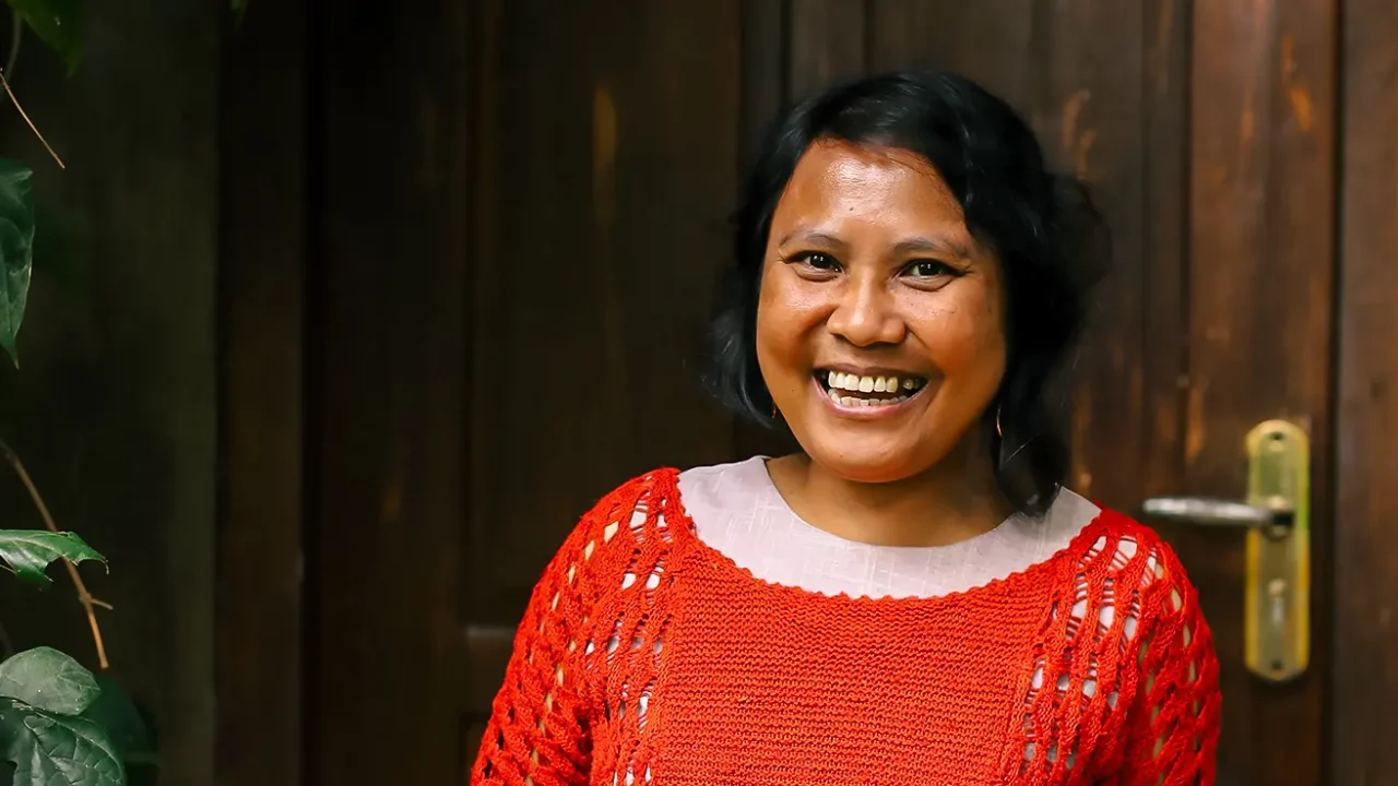 a woman, Lian Gogali, is wearing a red knitted sweater on top of white outfit while standing in front of a door.
