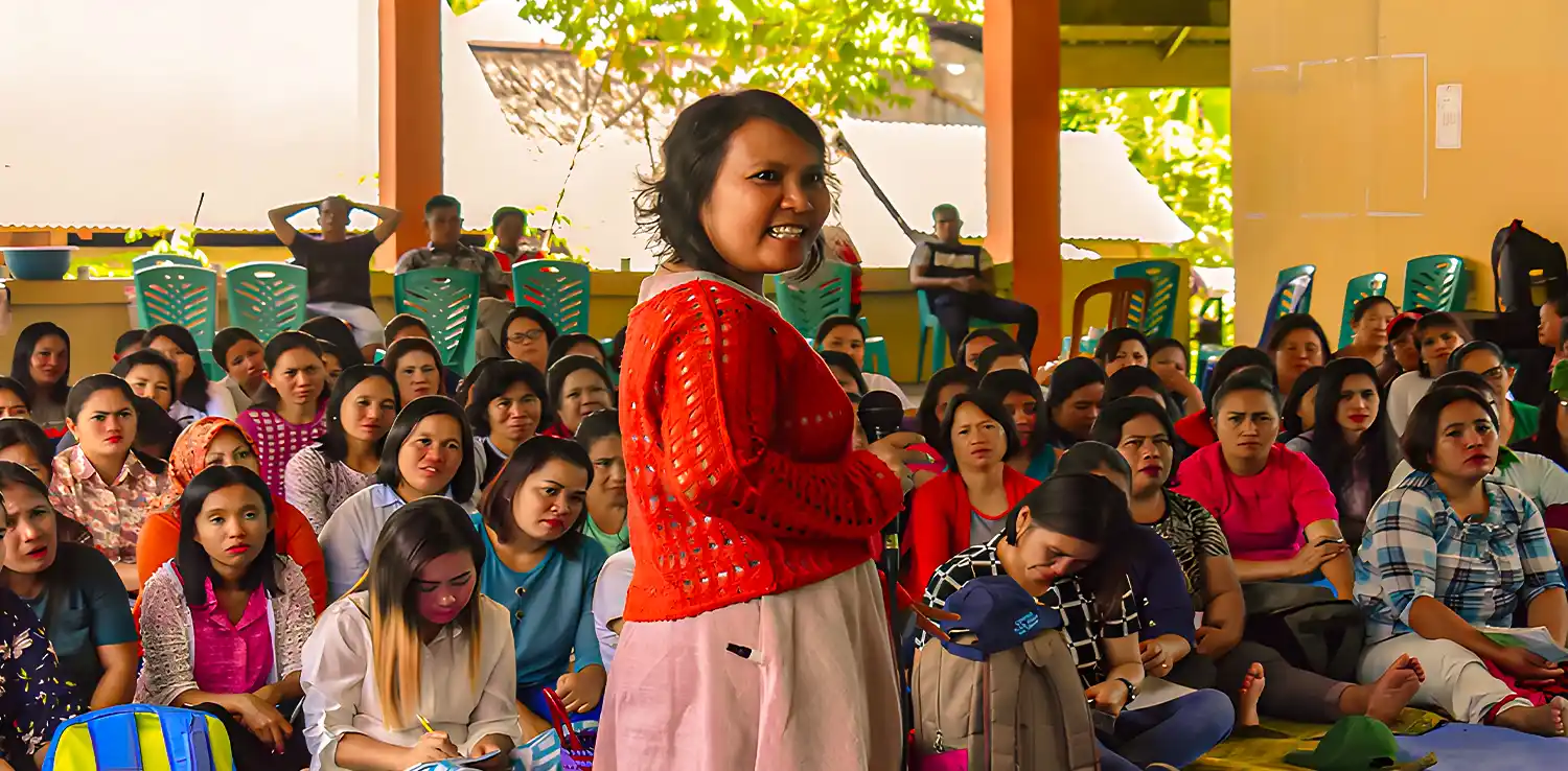 a woman, Lian Gogali, is standing in front of a group of women who are sitting on the floor