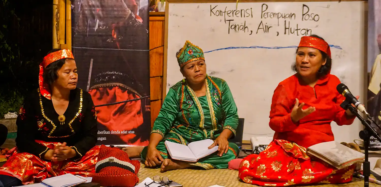 three women wearing Indigenous clothes are sitting on the floor in front of a white board.