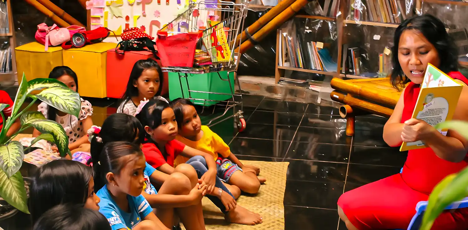 Lian Gogali showing a picture in a book to children who are sitting on the floor.