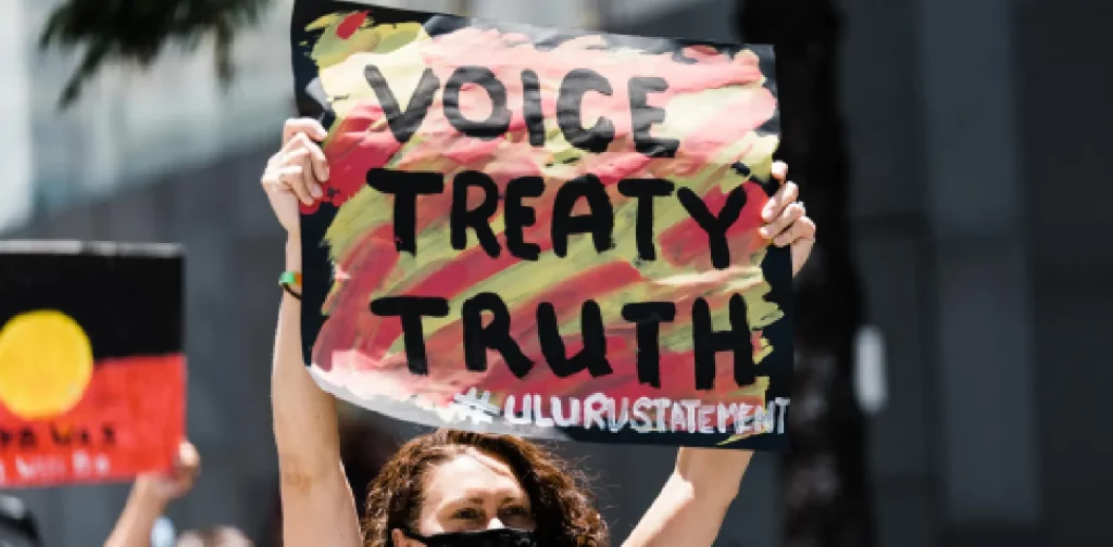 an indigenous Australian woman in mask showing a signage that says voice treaty truth