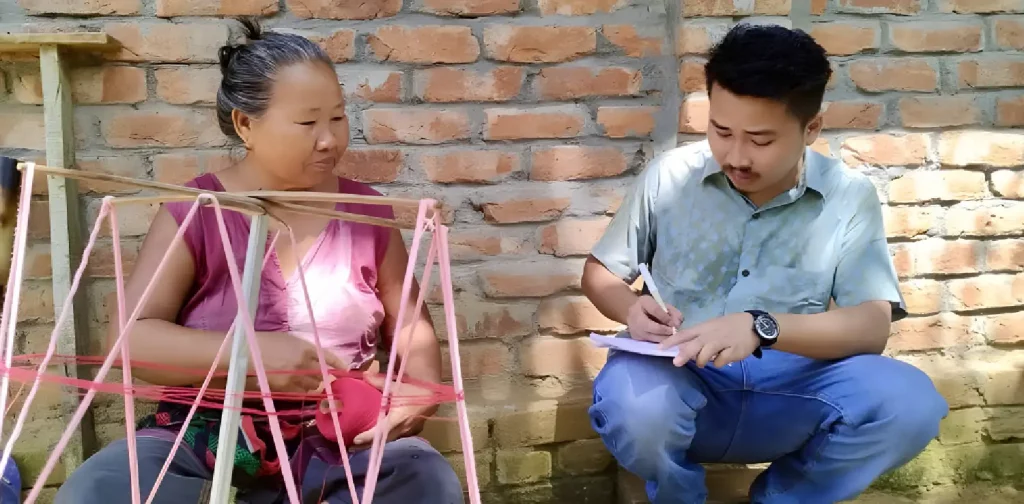 an elder lady sitting cross-legged and weaving talking to a young man crouching and writing on a note pad.