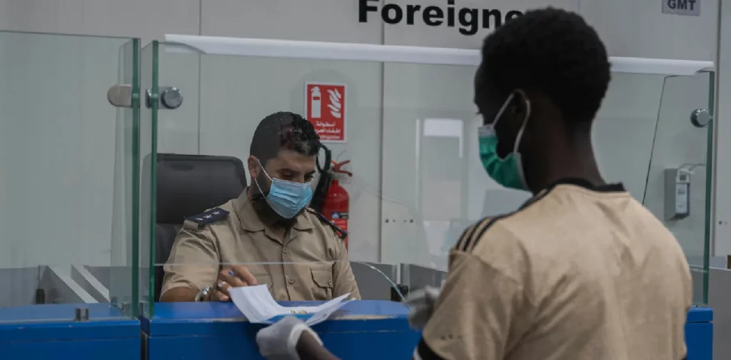 a man handing passport and migration documents to an officer behind a counter