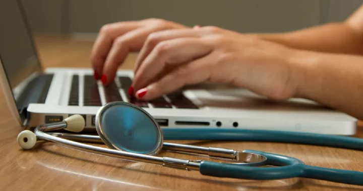 hands typing on a laptop with a stethoscope placed beside it