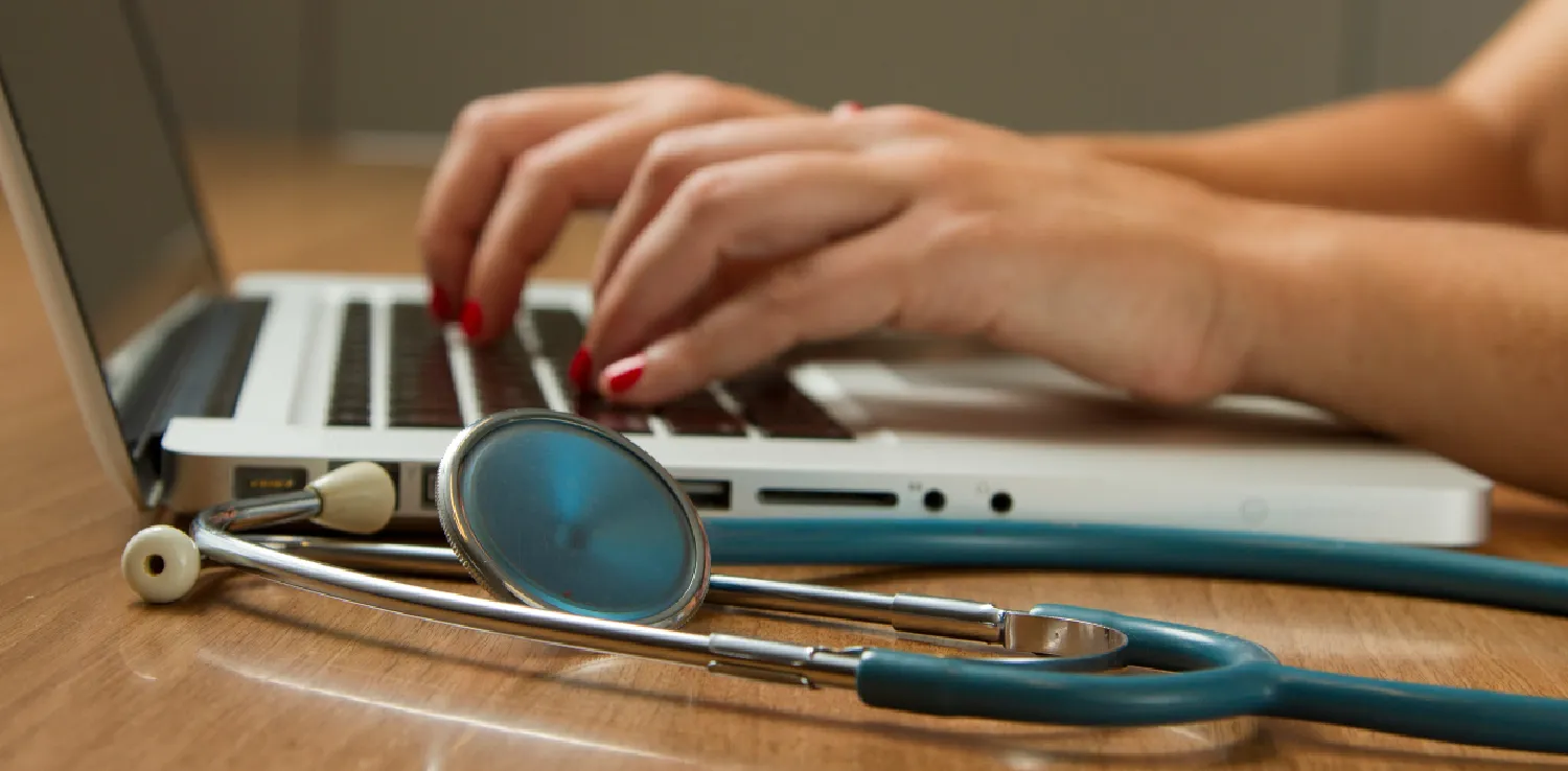 hands typing on a laptop with a stethoscope placed beside it