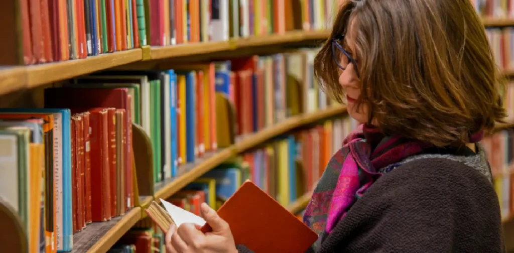 a woman with short hair and dark grey cardigan is reading a book taken from a shelf of a library