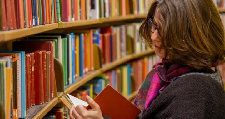 a woman with short hair and dark grey cardigan is reading a book taken from a shelf of a library