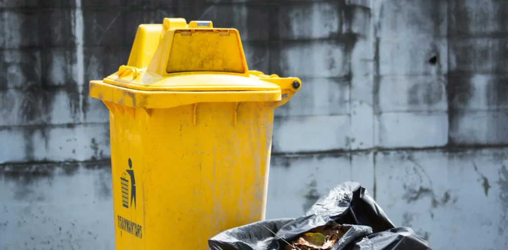 a yellow garbage bin with a plastic bag full of trash next to it