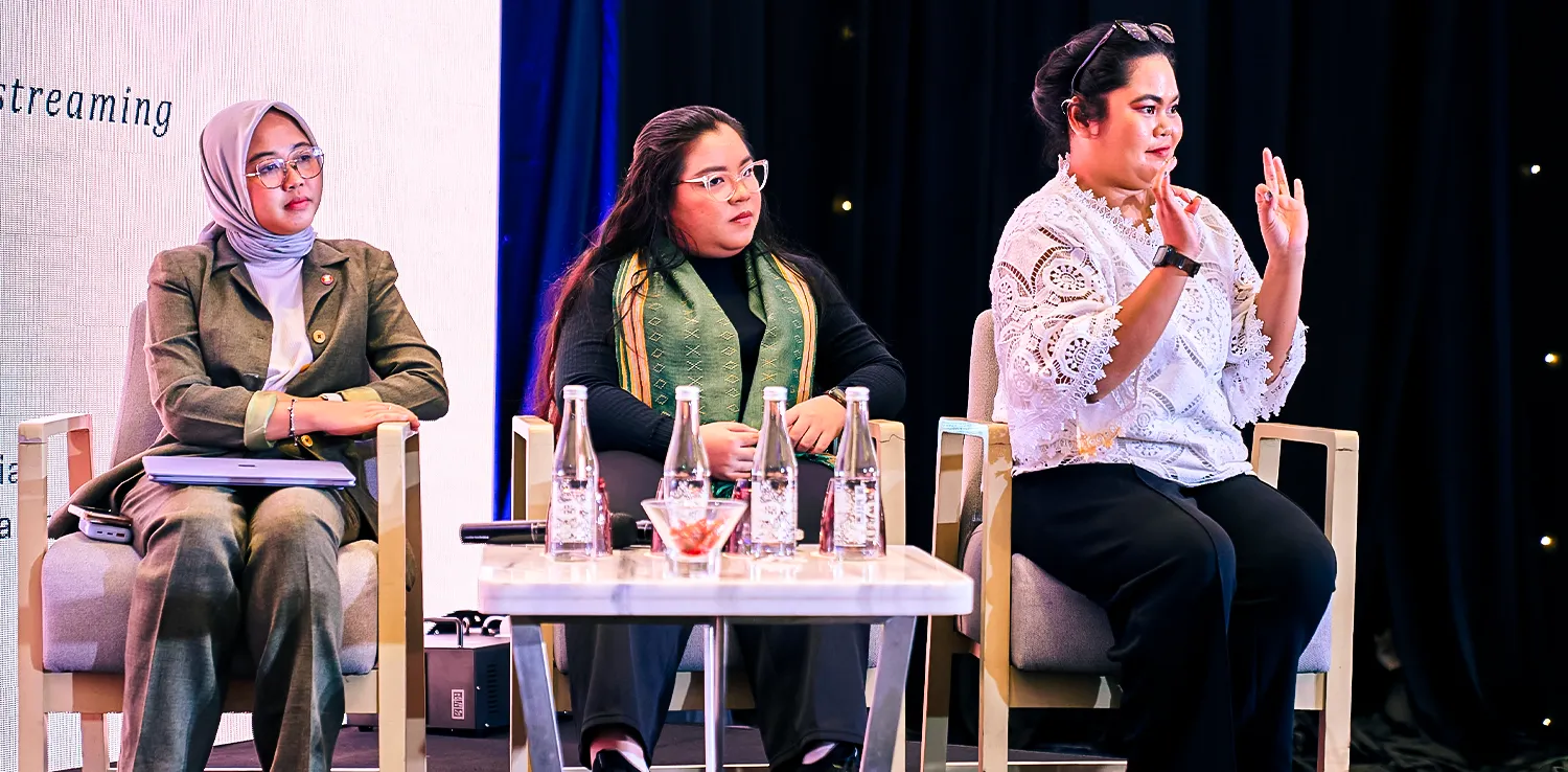 three women panelists sitting on stage, the one on the left wears hijab, the one in the middle wears glasses, and the one on the right is signing.