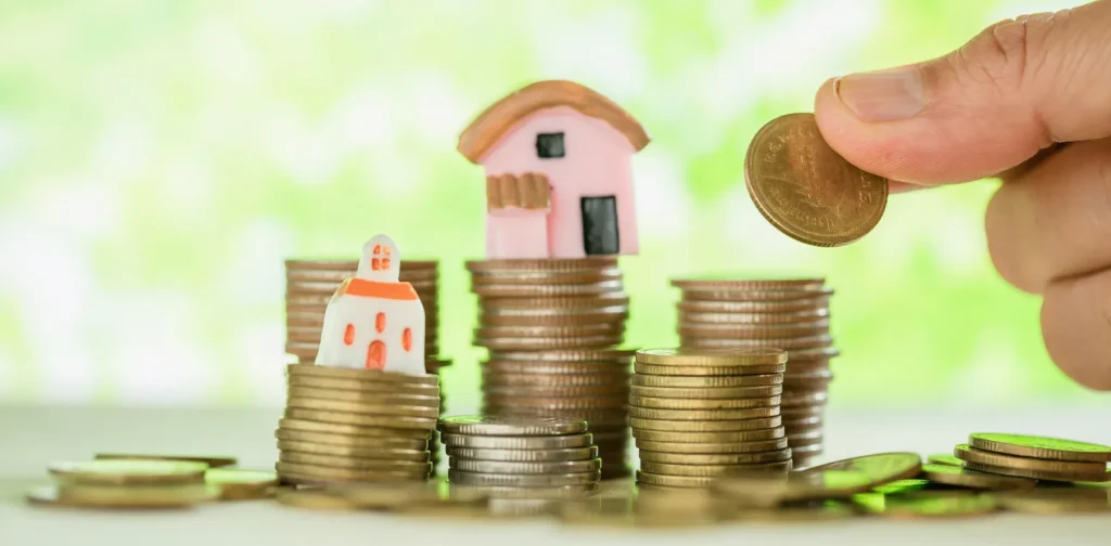 miniature of houses placed on top of coin piles, while a hand is adding coins to the pile