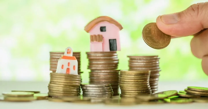 miniature of houses placed on top of coin piles, while a hand is adding coins to the pile