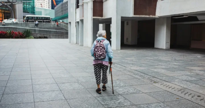 an elderly woman walking on a street with a walking stick, her back facing the camera