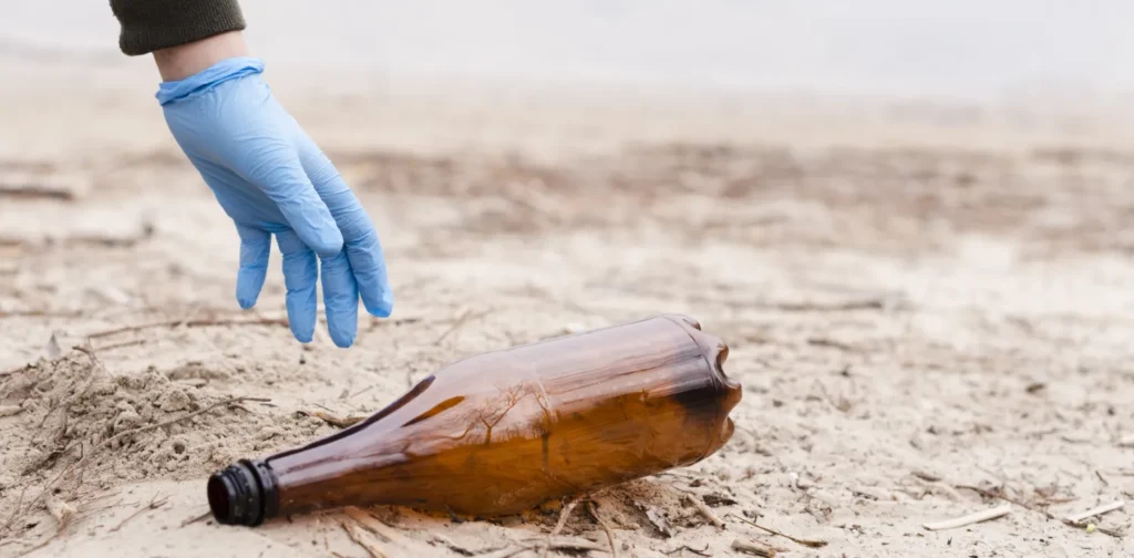 hand with blue glove picking up brown plastic bottle on sand