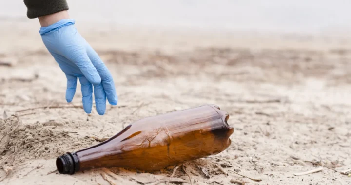 hand with blue glove picking up brown plastic bottle on sand