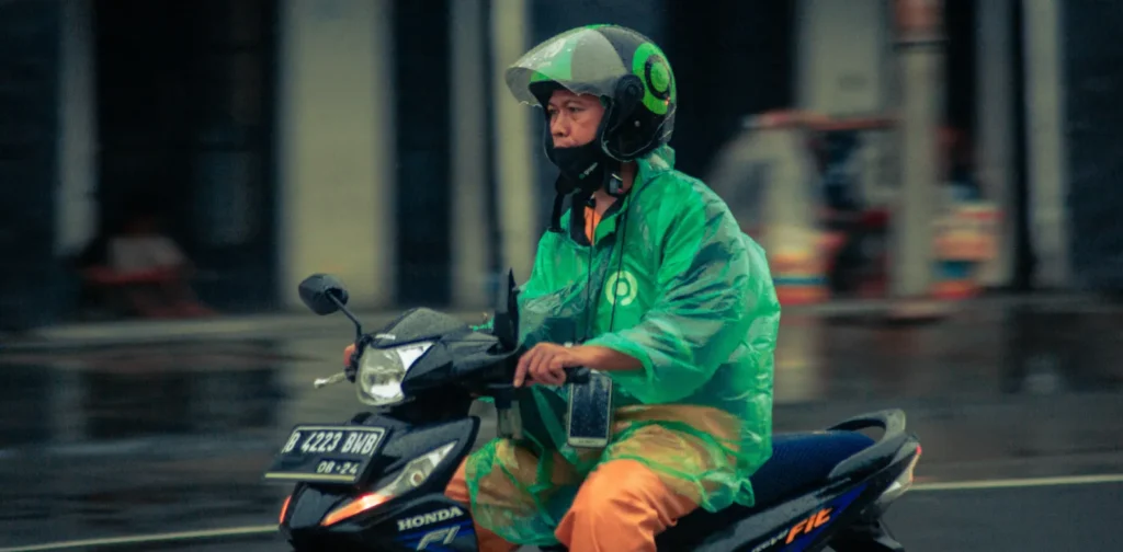 A man in a green jacket and green raincoat riding a black motorcycle on the road.