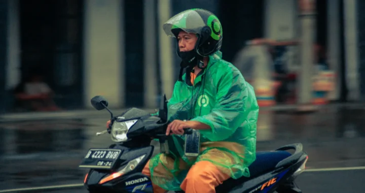 A man in a green jacket and green raincoat riding a black motorcycle on the road.