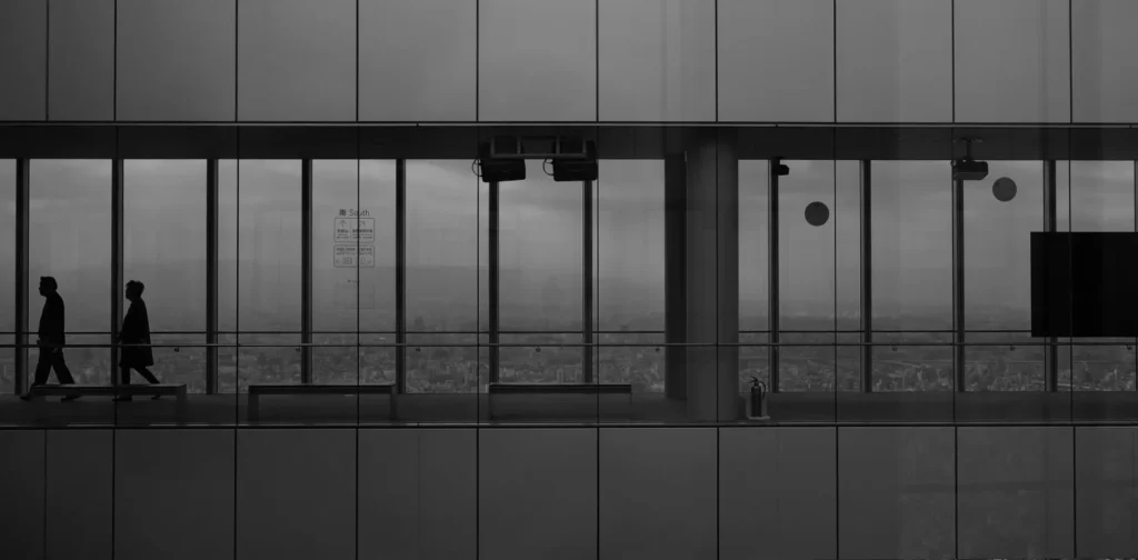 a grayscale photo of an office corridor with glass walls, with two men walking in it