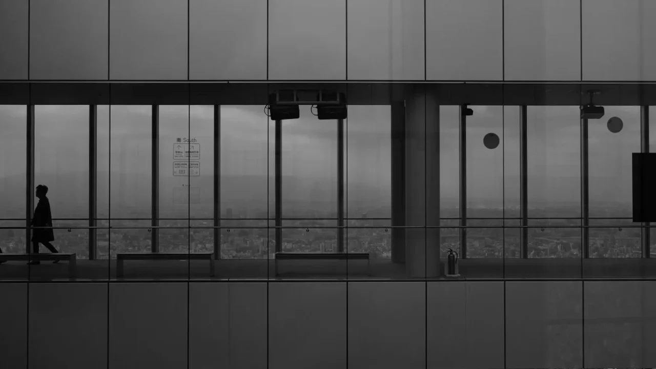 a grayscale photo of an office corridor with glass walls, with two men walking in it